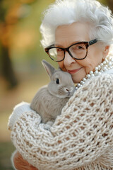 A smiling elderly woman with a stylish white hairdo embraces a grey rabbit against an autumnal background. scene is perfect for conveying the joy of pet ownership in later years.