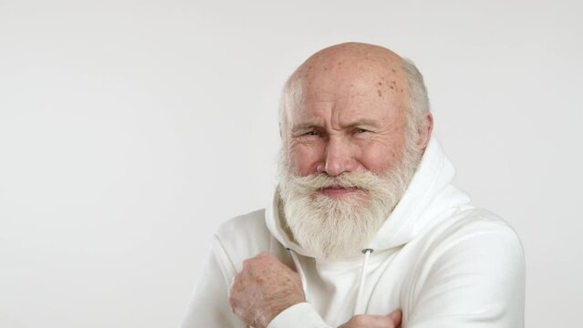 A sweet elderly man with a light beard in a white hoodie shows he's feeling cold by rubbing his hands together, set against a white background, depicting a chilly moment. Camera 8K RAW. 