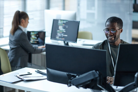Portrait of smiling Black man wearing glasses as IT programmer using computers at workplace in office copy space