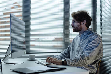 Graphic side view portrait of bearded man using computer in IT company office against windows with...