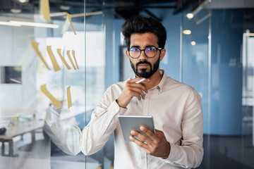 Young indian businessman contemplating during office work holding tablet - obrazy, fototapety, plakaty
