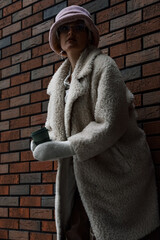 fashionable young beautiful woman in winter hat and fur coat with cup of coffee in her hands poses on background of brick wall on city streets in winter