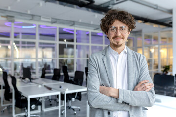 Portrait of a cheerful young businessman with curly hair confidently standing in a contemporary office space, arms crossed.