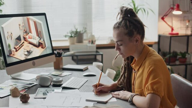 Waist up side view of young Caucasian female interior designer sitting at desk in creative workspace and drawing floor plan in sketchbook, looking at 3d rendering picture on desktop computer