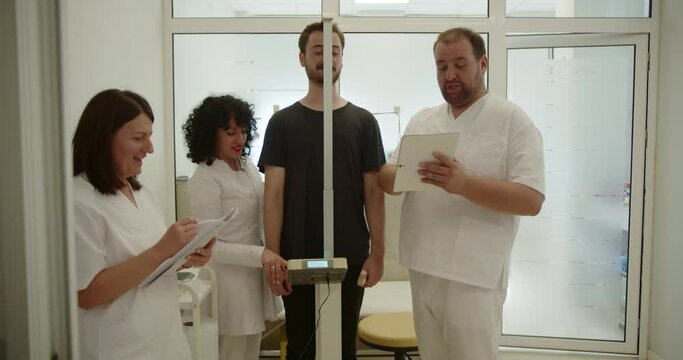 Three doctors measuring young patient height using stadiometer. They are having fun conversation and smiling while suddenly the patient read an unexpected news.