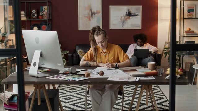 Dolly-in shot of Caucasian female interior designer looking at computer screen while drawing floor plan on sheet of paper sitting at desk in contemporary home office