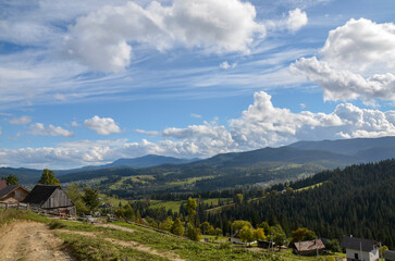 View of a village with wooden houses in a mountainous area surrounded by green meadows and mountains in the distance covered with dense forest under a clear sky with white clouds. Carpathians, Ukraine