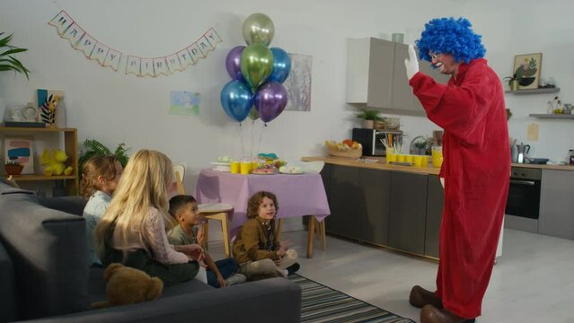 Side full footage of male clown in red costume and blue wig greeting children at birthday party in decorated living room at home