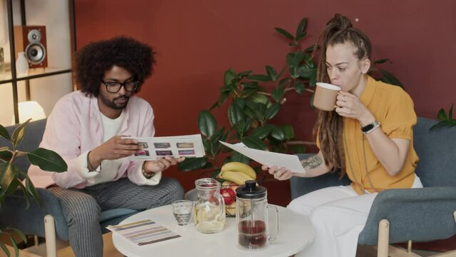 Medium shot of young creative ethnically diverse couple of interior designers sitting at coffee table in stylish office, chatting while looking at 3d rendering photos printed on sheets of paper