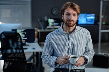 Waist up portrait of smiling bearded man holding coffee cup and looking at camera standing in...