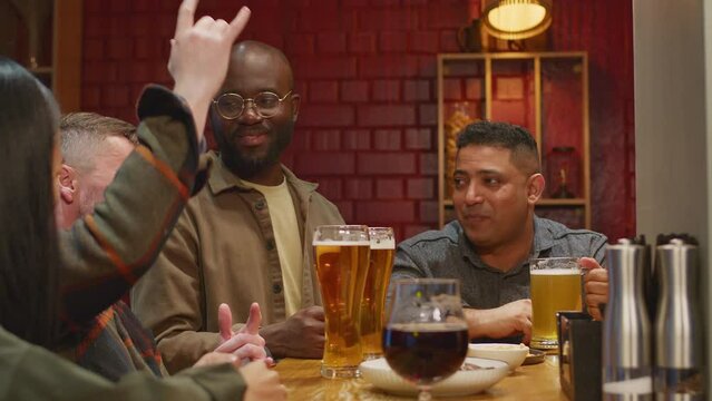 Side footage of group of diverse male and female adults hanging out in bar and celebrating clinking glasses of beer in evening