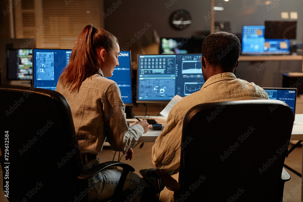 Wall mural back view at two it developers man and woman looking at computer screen with data in dark office
