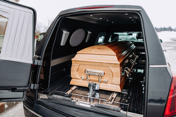 A coffin made of light wood stands in the trunk of a black hearse. Funeral and farewell ceremony. Closeup photo of a funeral casket