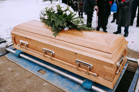 A wooden coffin with a bouquet of white flowers on the lid stands over a dug grave at the cemetery. Funeral ceremony at the cemetery. A funeral service and burial