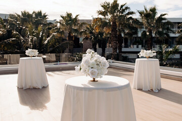 Elegant tables decorated with bouquets of white flowers, a beautiful festive setting outside for a wedding, festive event. Round tables with white tablecloths and bouquets of flowers without serving