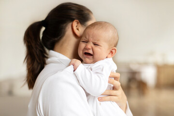 Young mother holding crying newborn baby girl or boy, calming kid, lullying in bedroom interior , focus on baby, free space