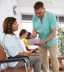 Obraz premium Middle-aged male medic and visitor sitting on chair discussing something written on paper in waiting hall