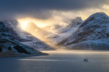 Snowy mountains in low clouds, bright sunbeams, boat in sea bay at colorful sunset in winter. Lofoten islands, Norway. Amazing landscape with rocks in snow, golden sun rays. North seaside. Nature - obrazy, fototapety, plakaty