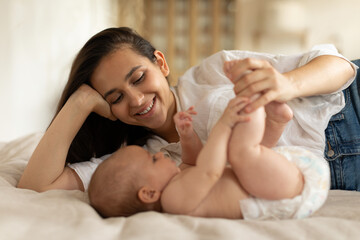 Happy motherhood. Young mother lying on bed with her small child girl, woman smiling, playing with baby daughter in bedroom at home