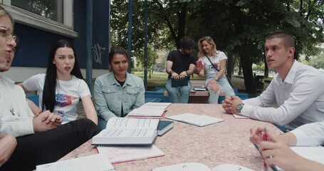 Diverse large group of students talking about the studies outside of the college campus sitting on a bench full of open books on a sunny day, trucking