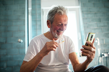 Smiling senior man brushing teeth and using smartphone in bathroom
