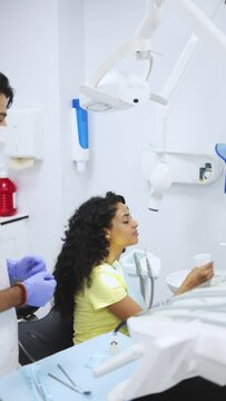 Vertical video of a young latin woman patient rinse her mouth over a dental cuspidor while her dentist is waiting to continue procedure. Young female patient rinses her mouth after treatment