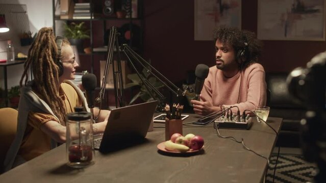 Waist up of young ethnically diverse man and woman sitting at table in dark studio, talking and recording video podcastWaist up of young ethnically diverse man and woman sitting at table in dark studi