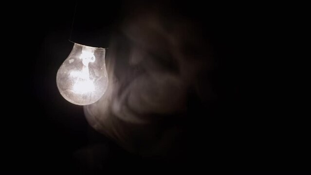 One Hanging, Glowing Light Bulb in Thick Smoke on a Black Background. Swinging, Dusty, switched-on Edison tungsten light bulb shines a yellow light in a smoky dark room. Blurred motion. Texture. Fog.