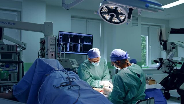 Doctors bent above the patient's arm operating the hand. Rear view of a surgeon sitting at operational table wearing loupes.