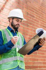 Latin construction worker man looking documents with  brick wall at background in sunny day