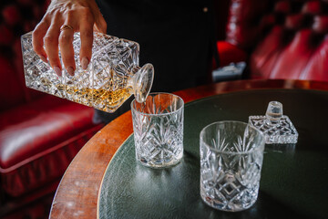 Valmiera, Latvia - July 7, 2023 - A hand is pouring whiskey into a glass from a crystal decanter on a table.