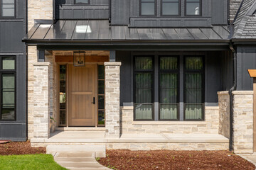 A front door detail on a home with black board and batten siding with natural stone accents and a beautiful oak front door.  - obrazy, fototapety, plakaty
