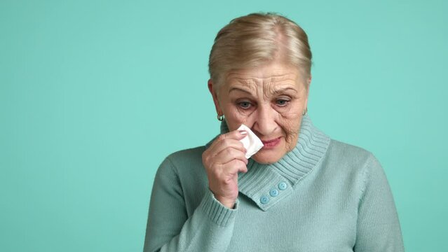 Old woman in grief sobbing, female pensioner of 60-70 years with blonde short hair in blue sweater standing over blue background crying, gulping down sobs. High quality 4k footage