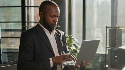 Fototapeta premium African American multiethnic businessman employee professional manager using laptop typing email on keyboard focused man search digital data standing with computer in modern office sunbeam background