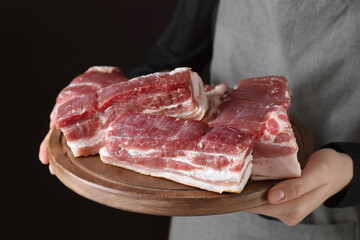 Woman holding wooden board with pieces of raw pork belly on black background, closeup