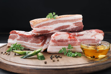 Pieces of raw pork belly, chili pepper, peppercorns, oil and parsley on black wooden table, closeup