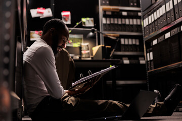 African american policeman reading crime case folder on floor in dark office. Detective examining...