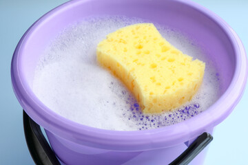 Bucket with foam and sponge on light blue background, closeup