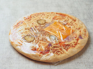 Burger style Italian pizza with tomato sauce, meat, cucumber and cheese and special sauce in plastic wrap on a light color table cloth. Ready meals product for cooking home.