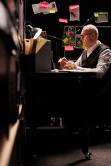 Elderly man police officer sitting at desk table in evidence room, working overtime at missing person case. Senior private investigator analyzing victim files, looking at crime scene report