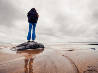 Teenager girl in black jacket standing on a rock at sandy beach, cloudy sky. Lahinch, county Clare,...
