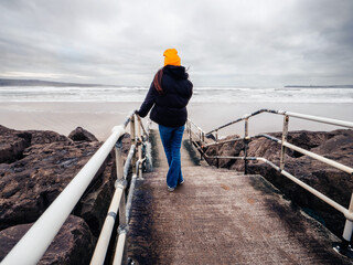 Teenager girl in black jacket standing on a footpath to a vast sandy beach and ocean, dramatic sky in the background. Trip to nature concept. Lahinch, county Clare, Ireland. Travel and tourism theme. - obrazy, fototapety, plakaty