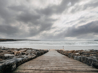 Fototapeta na wymiar Ramp to a vast sandy beach and rough ocean water, silhouette of a person walking along shows the scale of the place. Lahinch, county Clare, Ireland. Dark dramatic sky. Rough nature scene.