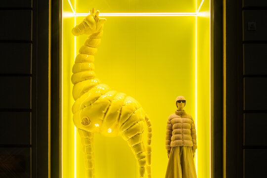 Florence, Italy - August 29 2023 : A stylish window display for luxury brand moncler with giraffe in puffer coat and manikin in coat, glasses and skirt at night. Bright neon yellow and white light