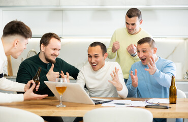 Group of men watching sports match online on laptop during friendly meeting with beer at home, excitedly celebrating winning score in game ..