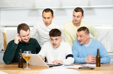 Fototapeta premium Focused group of male friends gathering around laptop, discussing and planning event with drinks on table at home..
