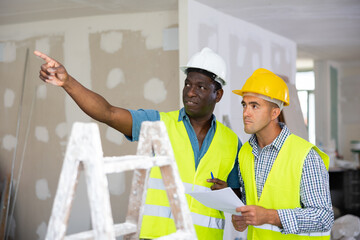 Foreman and worker in yellow vest and protective helmet are discussing the repair of a room in a cottage