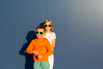 Eight-year-old boy and girl in colorful bright sunglasses playing and laughing on the street against the background of a blue wall - 729673104
