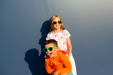 Eight-year-old boy and girl in colorful bright sunglasses play and she tickling him on the street against the background of a blue wall - 729673113