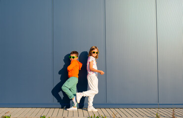 Eight-year-old boy and girl stand with their backs to each other on the street against the background of a blue wall - 729673127